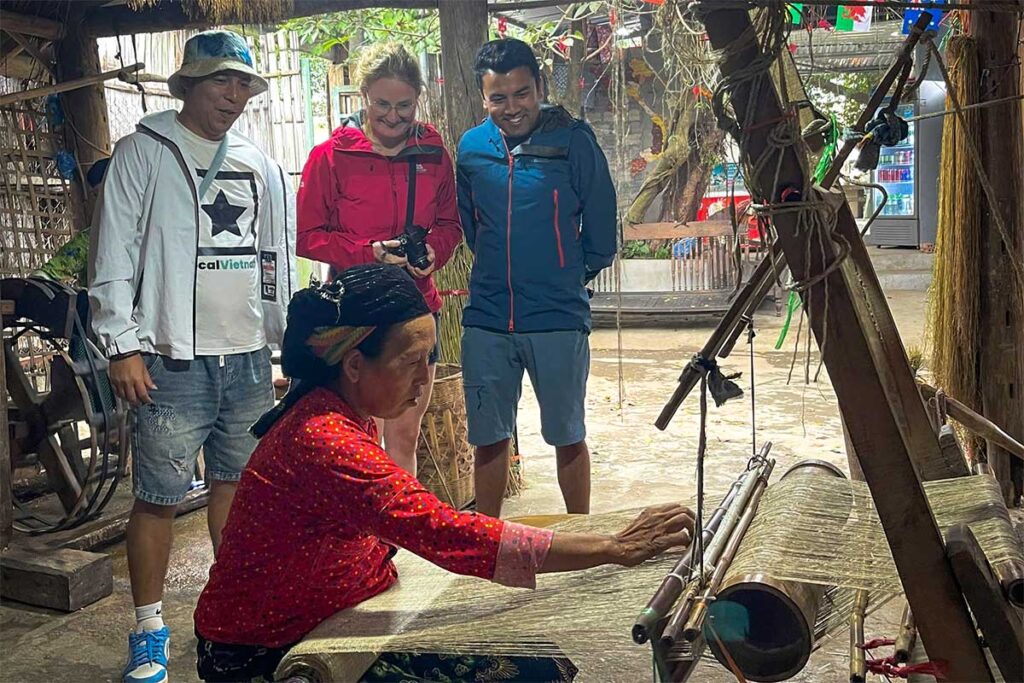 Local guide and travelers observing traditional hemp weaving in Lung Tam village on a Ha Giang Loop tour, showing authentic Hmong textile craftsmanship