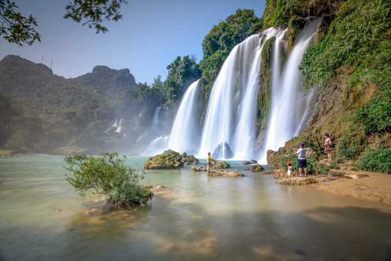 Most beautiful nature places in Vietnam, featuring Ban Gioc Waterfall cascading into a turquoise river surrounded by lush jungle and limestone hills.
