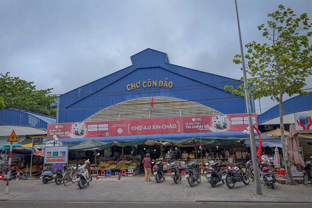 Main entrance of Con Dao Market with motorbikes parked outside and vendors selling goods in front of the market hall