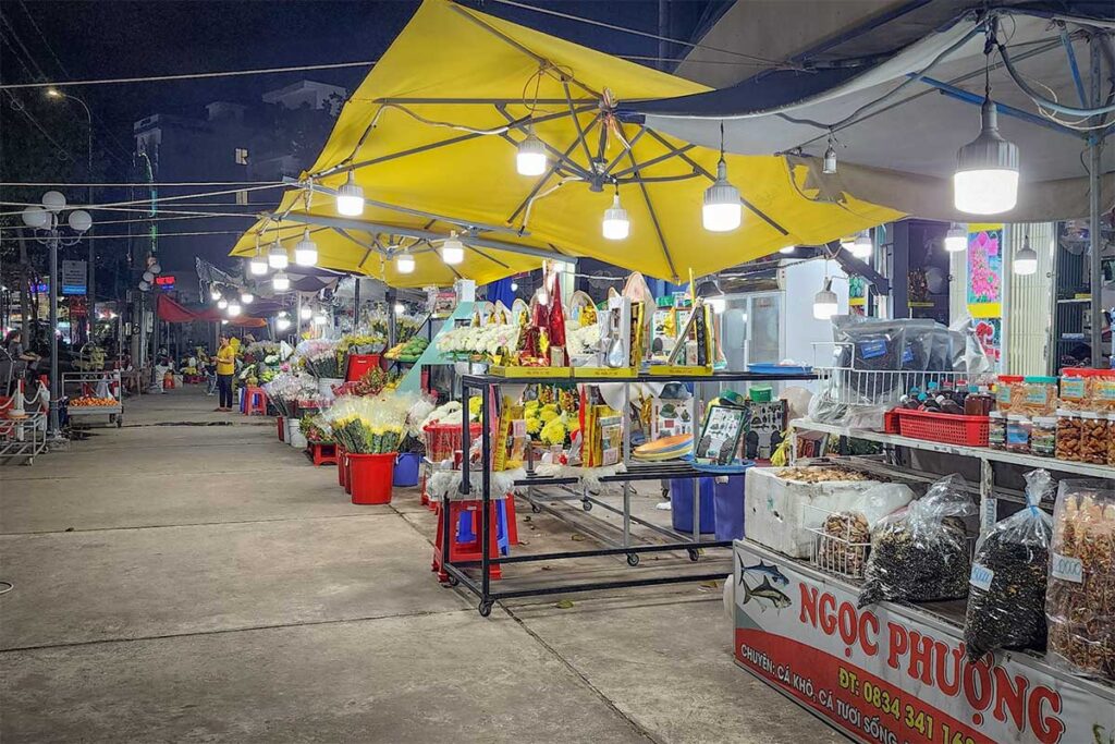 Night market stalls in Con Dao selling flowers, incense and offerings alongside snacks and souvenirs under bright lights in the evening