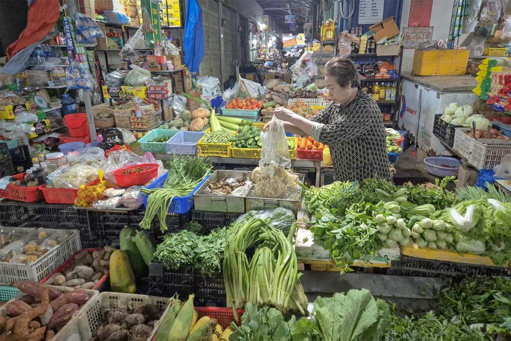 Vegetable stall inside Con Dao Market with a local vendor preparing fresh produce, including leafy greens, herbs, and root vegetables in a traditional indoor market setting