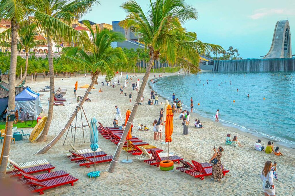 Busy beach in Sunset Town Phu Quoc with palm trees sunbeds people relaxing and modern waterfront architecture