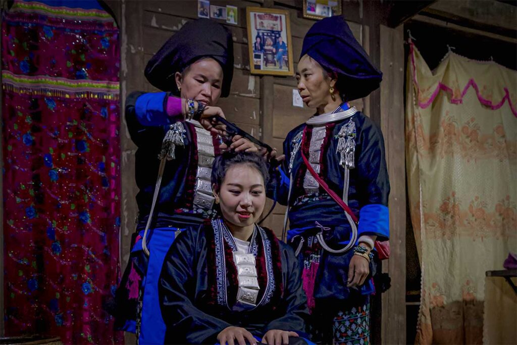 Local women in traditional ethnic clothing preparing a hairstyle inside a wooden house in Sin Ho, showing cultural customs and daily life