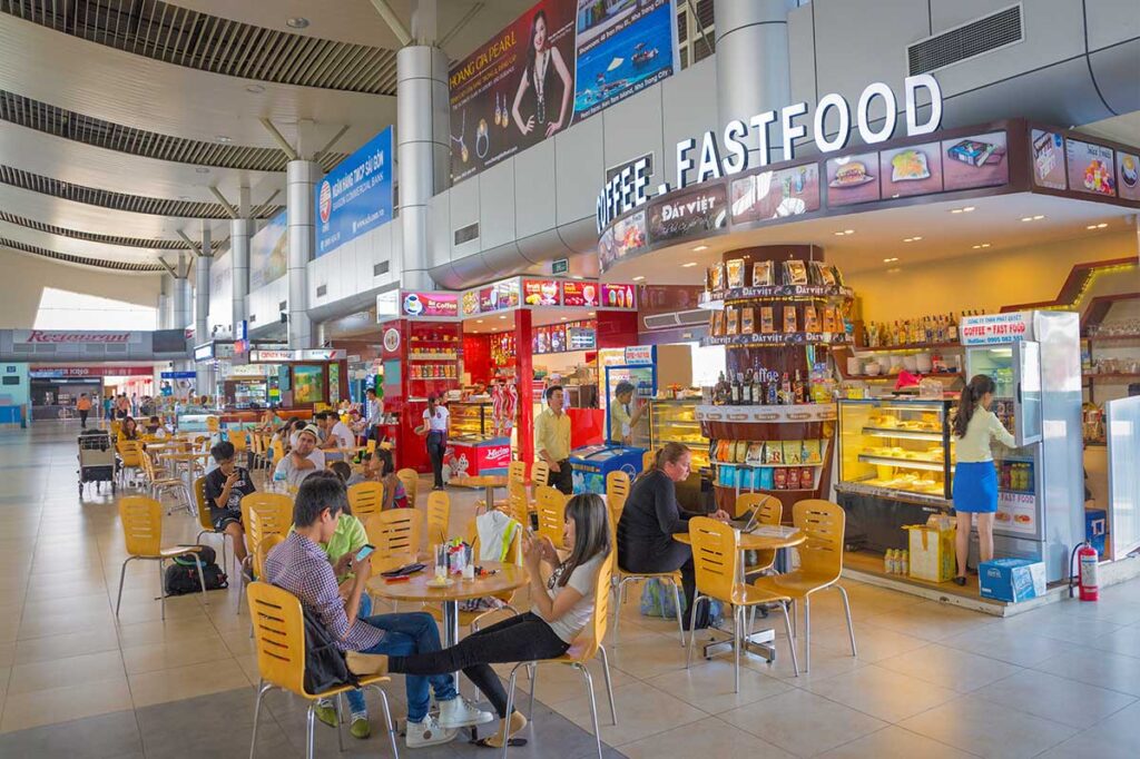 Interior of Cam Ranh Airport showing café seating area with passengers, food stalls and drink counters in the terminal