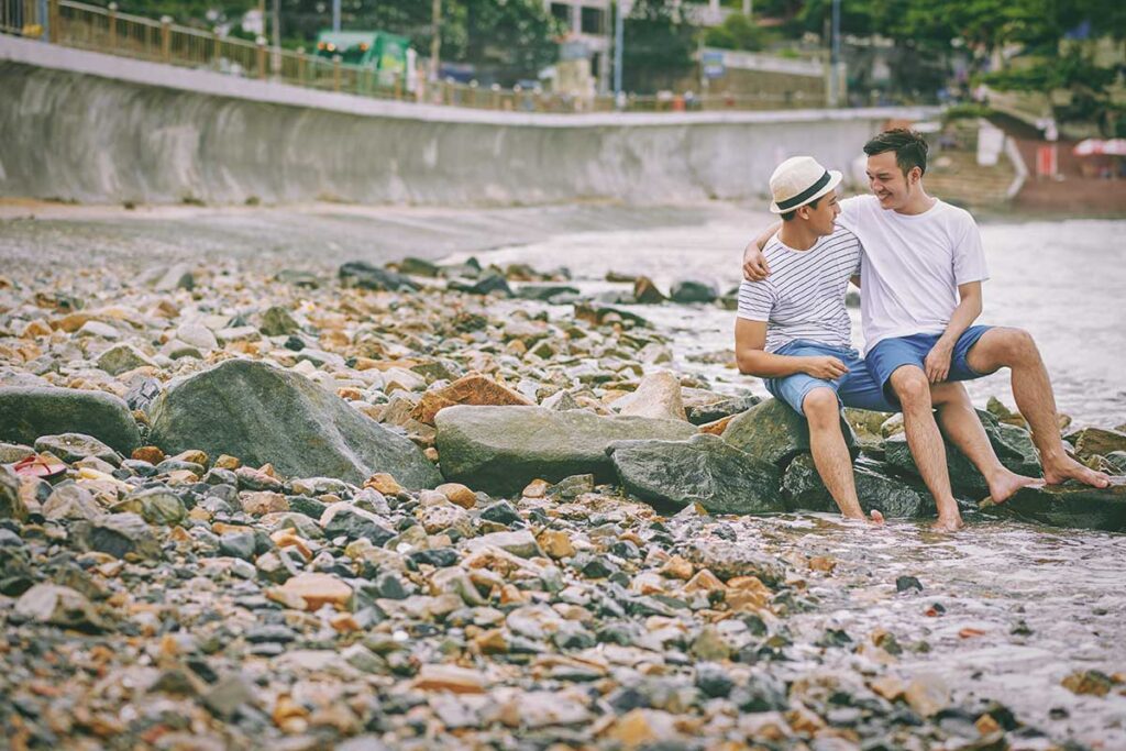 gay couple sitting together on rocks by the sea in Vietnam showing relaxed and safe LGBTQ travel atmosphere