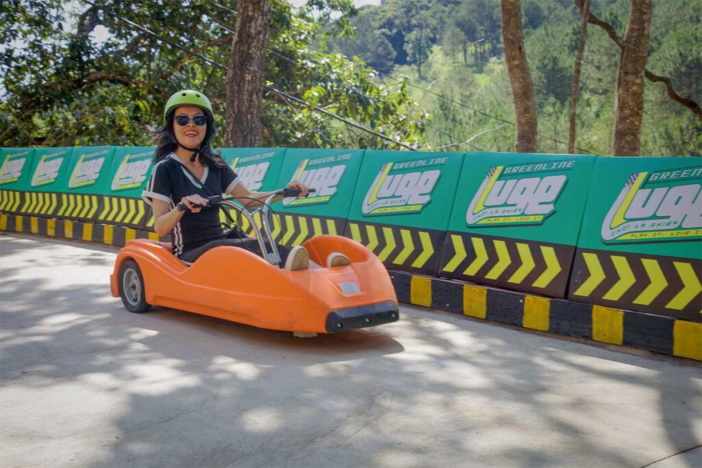 Person riding a downhill luge cart on the Mario Kart Dalat track, wearing a helmet and driving through a curved section of the course