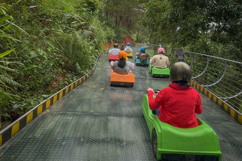 Group of visitors riding downhill luge carts on the Mario Kart Dalat track, following a curved section through forest scenery at Cao Nguyen Hoa
