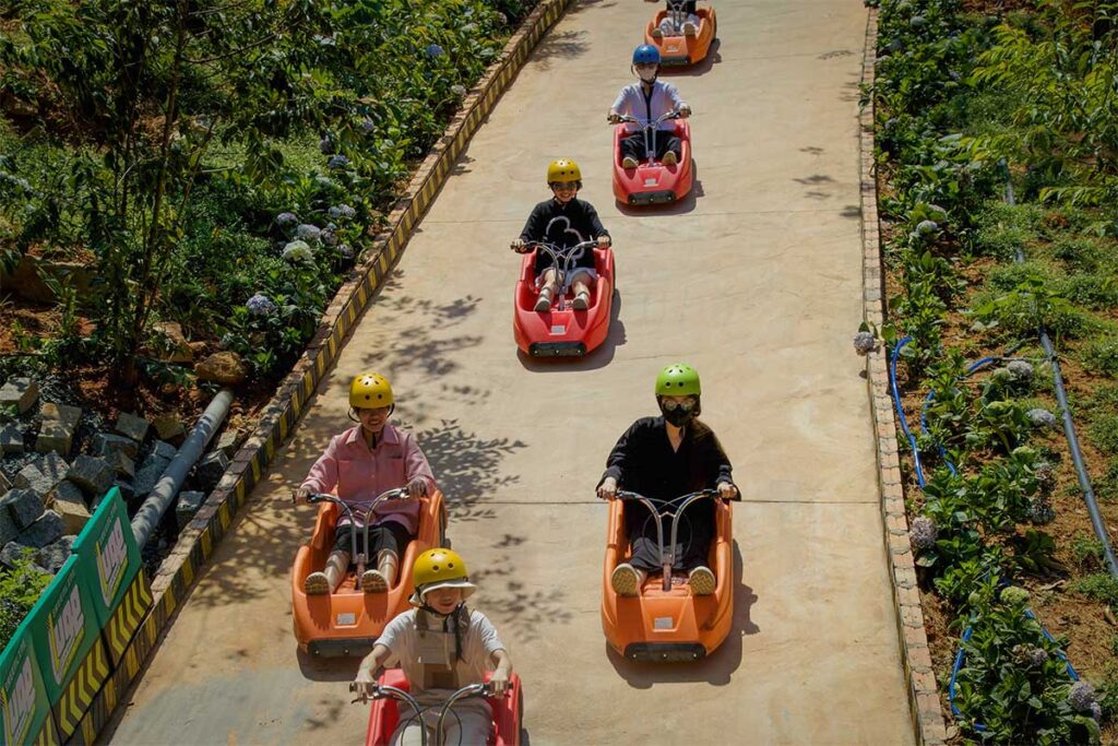 People riding colorful luge carts downhill at Mario Kart Dalat, spaced along the track with views of the surrounding gardens