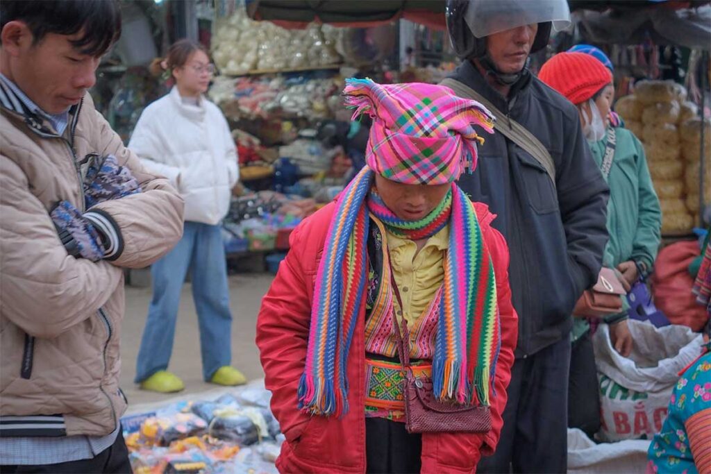 Ethnic minority woman wearing colorful traditional clothing at a local market in Sin Ho, Lai Chau, surrounded by stalls and everyday trading activity