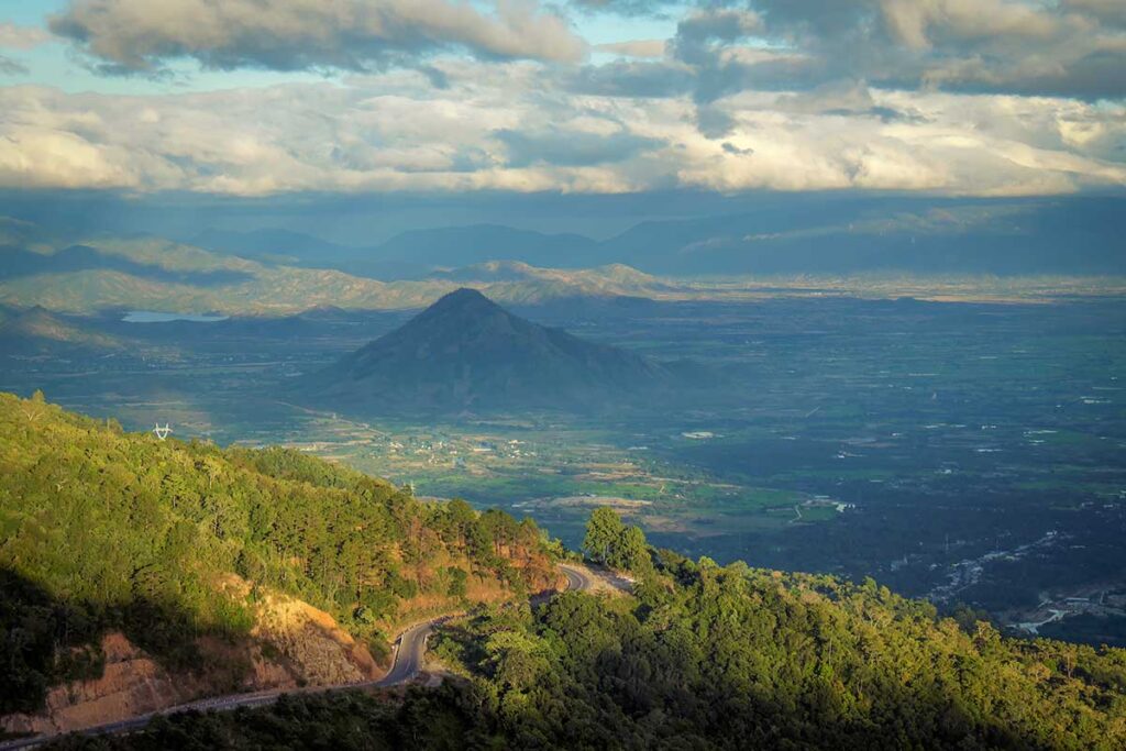 Wide view over Ngoan Muc Pass with winding mountain road descending toward the Phan Rang plains, surrounded by green hills and layered mountains in the distance