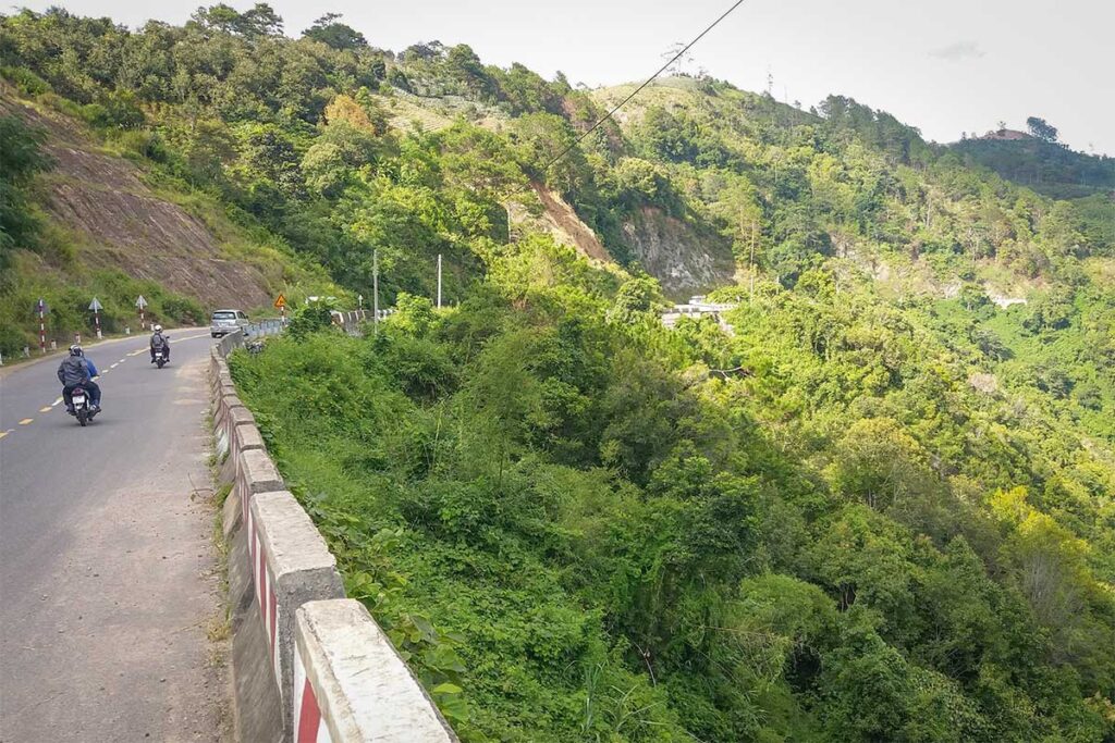 Motorbikes riding along a curved section of Ngoan Muc Pass with lush green hills and steep mountain slopes on both sides