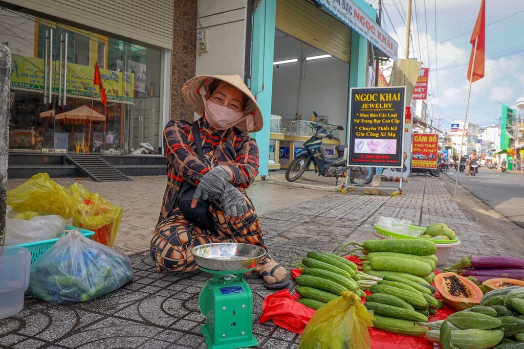 local street vendor in Vietnam wearing a mask while selling vegetables along a busy road