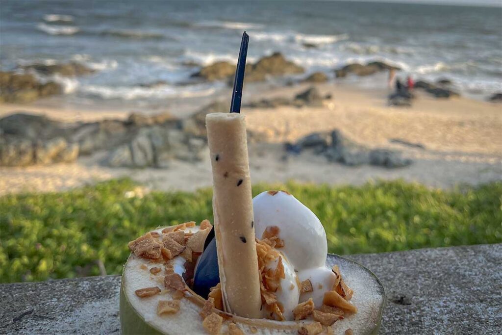 Coconut ice cream served in a fresh coconut shell at Ong Dia Beach in Mui Ne, with the rocky shoreline and ocean waves visible in the background.