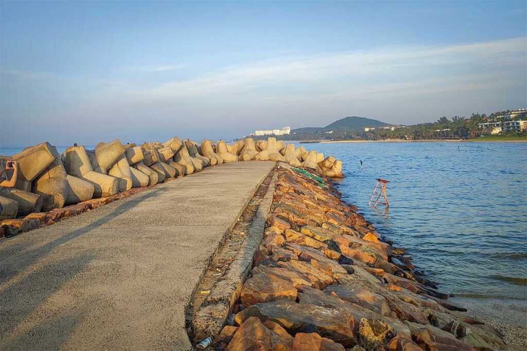 The concrete walkway along the breakwater at Ong Dia Beach in Mui Ne at golden hour, flanked by large concrete blocks on one side and rocks leading into the calm sea on the other, with the coastline and a hill visible in the distance.