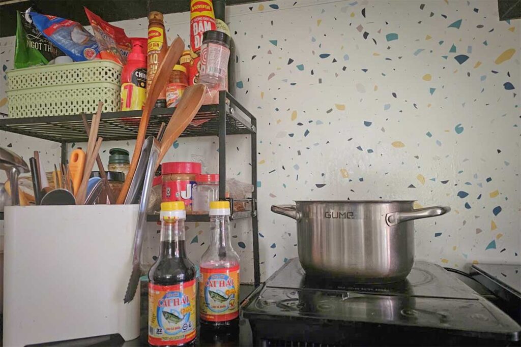 Two bottles of Cat Hai fish sauce on the counter of a typical Vietnamese home kitchen, next to a cooking pot on the stove and a shelf stocked with condiments and utensils.