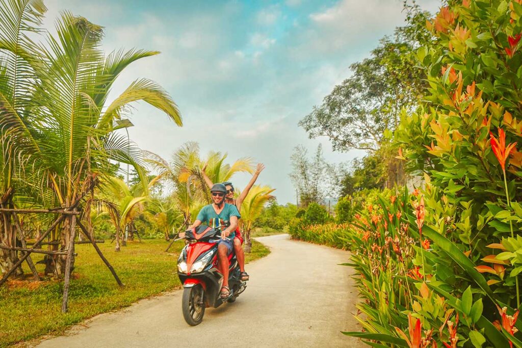 Couple riding a rental scooter through a tropical road in Phu Quoc, surrounded by palm trees and lush vegetation
