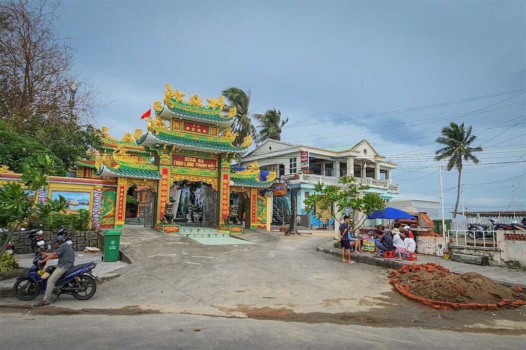 Street view of Dinh Ba Thuy Long Thanh Mau Temple in Phu Quoc showing the temple entrance small local houses motorbikes and daily life around the area