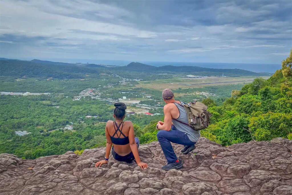 Two hikers sitting on rocky boulders at the summit of Dinh Tien Son Peak in Phu Quoc, with panoramic views over the island and the international airport below