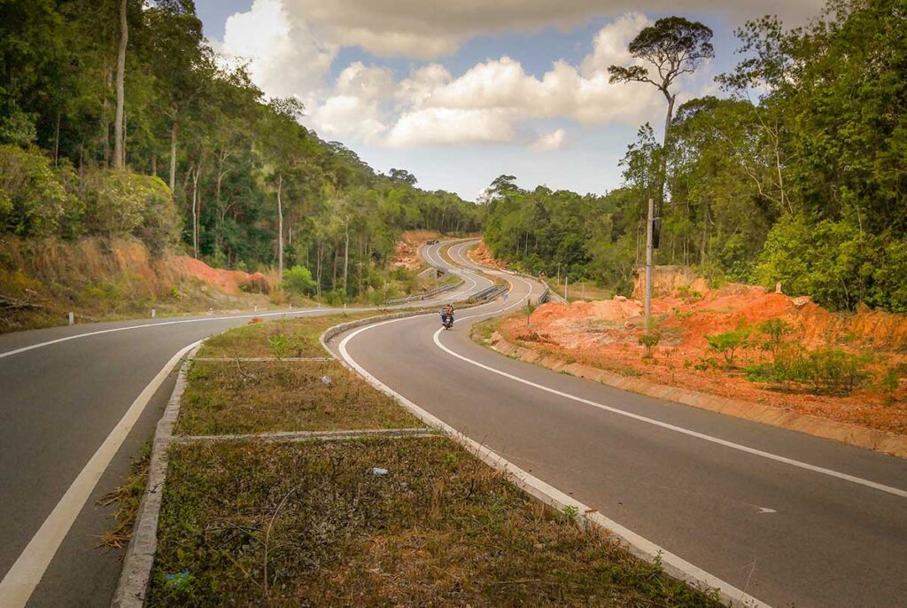 Well-paved winding road through the jungle in Phu Quoc, with a motorbike rider navigating a long curve between forested hills