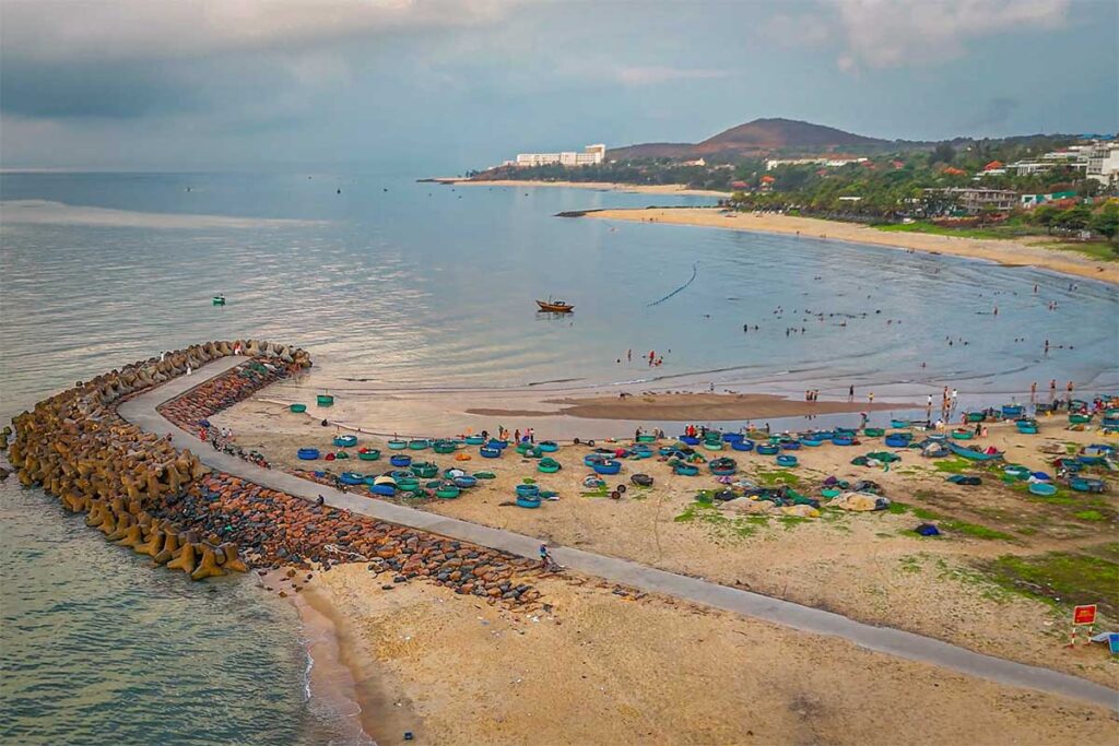 Drone view of Ong Dia Beach in Mui Ne showing the curved concrete breakwater, dozens of basket boats on the sandy shore, swimmers in the calm water, and the coastline stretching toward Phan Thiet in the background.