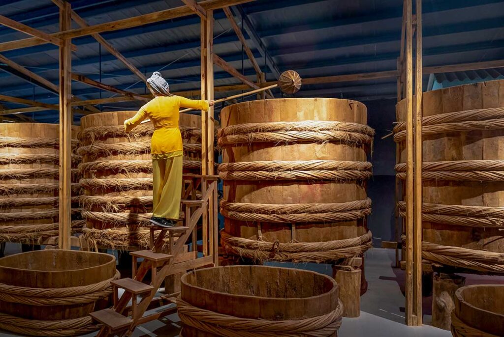 Traditional fish sauce fermentation room at Lang Chai Xua Fish Sauce Museum with large wooden barrels and a costumed worker demonstrating the brewing process