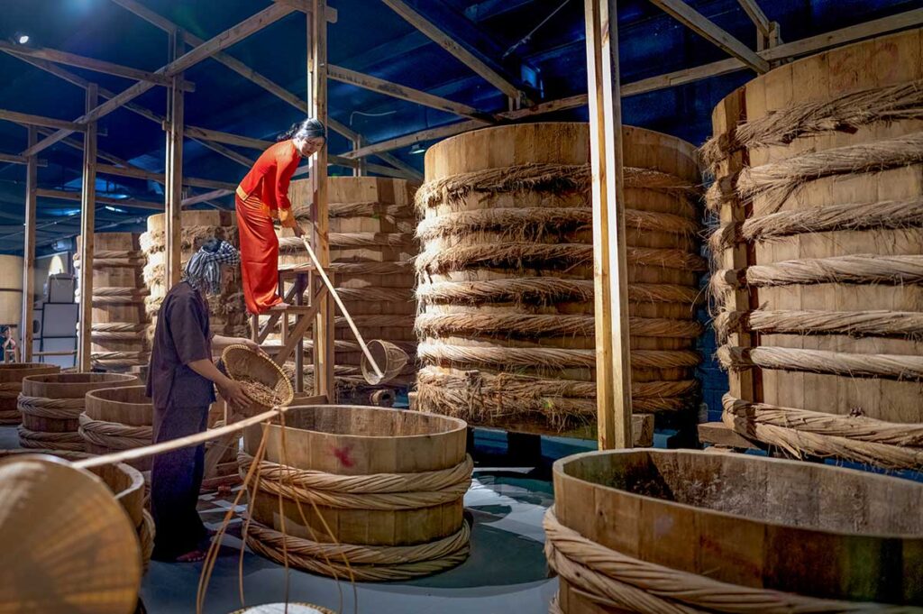 Two costumed workers demonstrating the traditional fish sauce fermentation process among large wooden vats inside Lang Chai Xua Fish Sauce Museum in Phan Thiet