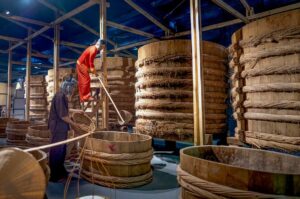 Two costumed workers demonstrating the traditional fish sauce fermentation process among large wooden vats inside Lang Chai Xua Fish Sauce Museum in Phan Thiet