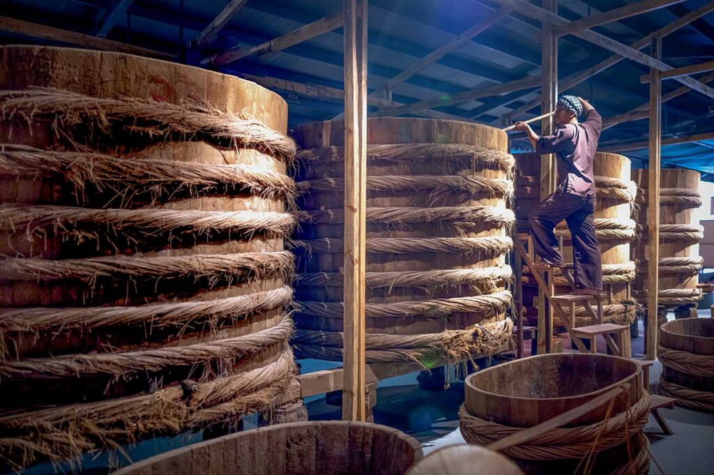 Life-size reconstruction of traditional fish sauce fermentation vats inside Lang Chai Xua Fish Sauce Museum, with a worker figure climbing a wooden ladder to tend the barrels