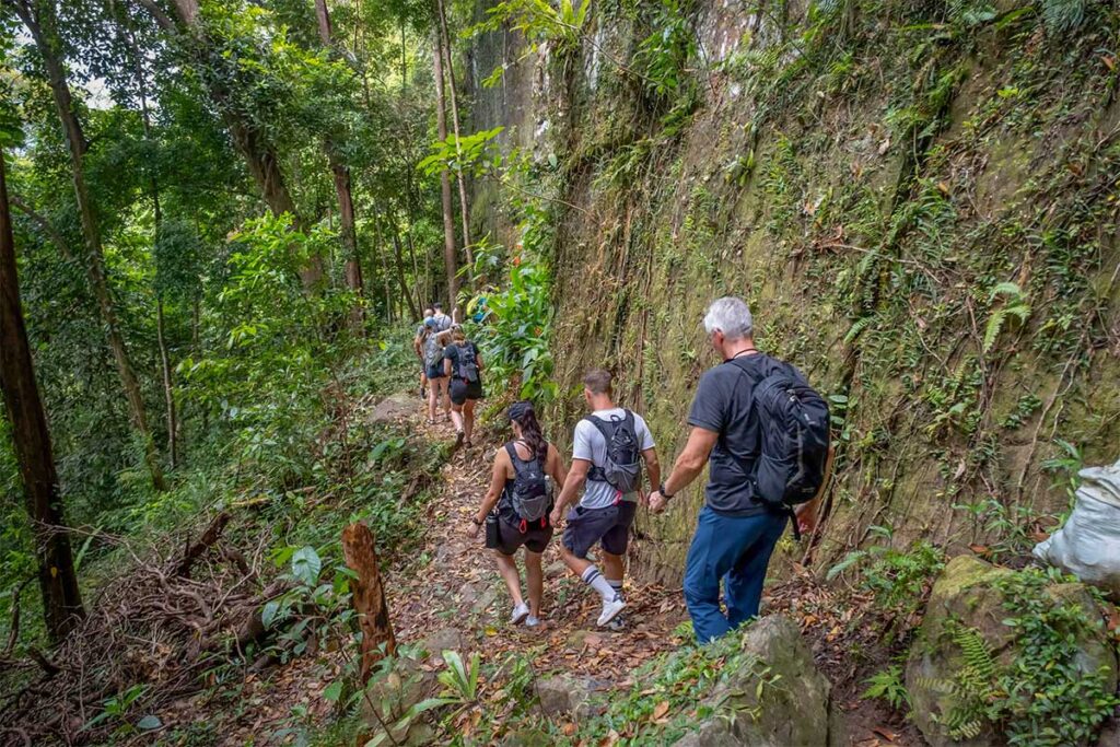Group of hikers walking a narrow jungle trail alongside a moss-covered rock face during a guided trekking tour to Dinh Tien Son Peak in Phu Quoc