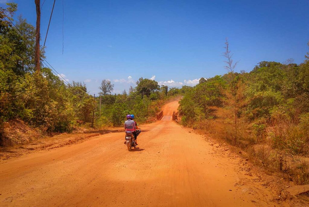 Two riders on a motorbike navigating a red dirt road in the north of Phu Quoc, flanked by dry vegetation under a clear blue sky