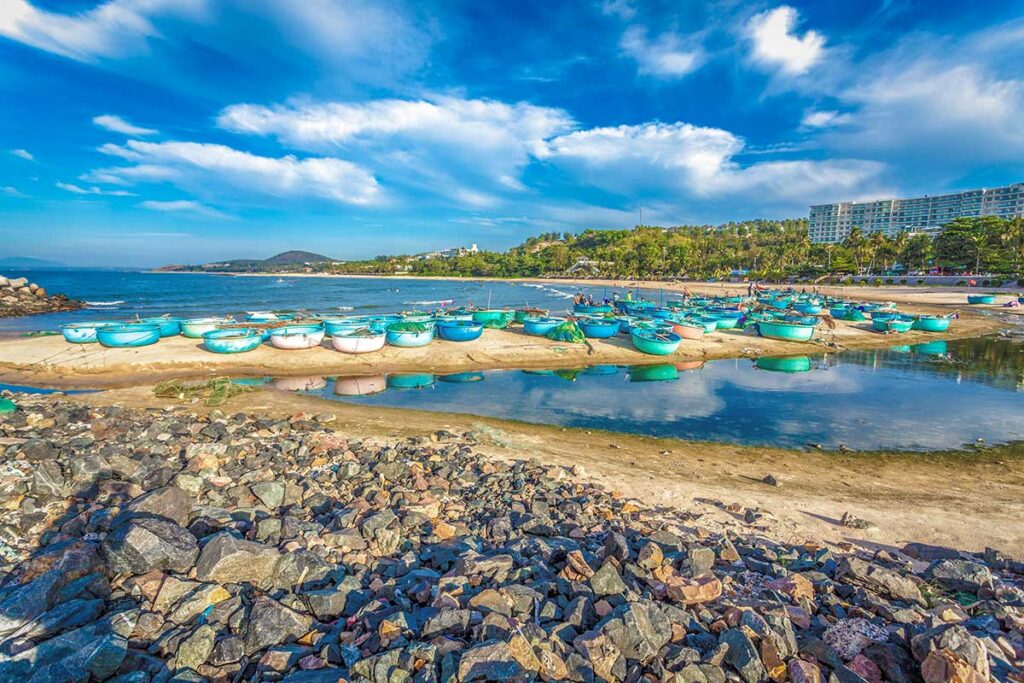 Rows of turquoise basket boats lined up along the sandy shore at Ong Dia Beach in Mui Ne, with the rocky breakwater in the foreground and a resort building visible among the trees in the background.