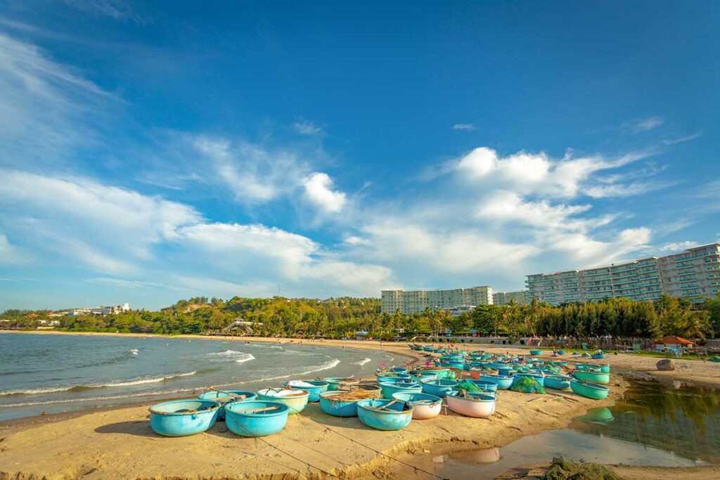 Dozens of traditional round basket boats resting on the sand at Ong Dia Beach in Mui Ne on a sunny day, with the beach and resort buildings stretching along the coastline in the background.