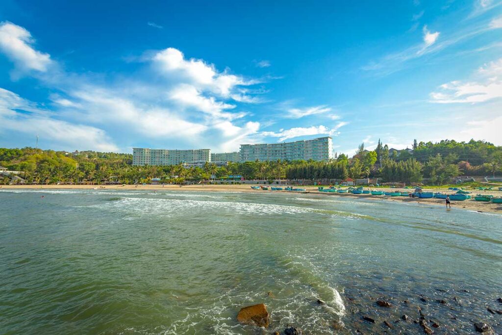View of Ong Dia Beach in Mui Ne from the water, showing waves rolling in toward the sandy shore, a row of basket boats pulled up on the beach, and a large resort building rising above the palm trees in the background.