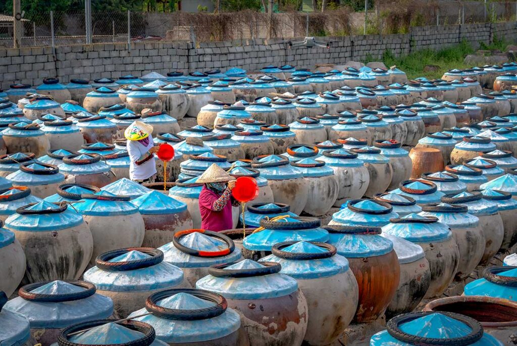 Two workers in traditional Vietnamese conical hats pouring fish sauce between large ceramic fermentation jars at an outdoor Phan Thiet fish sauce factory.