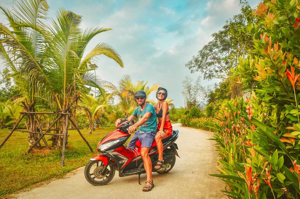 Two tourists on a rental scooter posing on a quiet tropical road in Phu Quoc, helmets on, surrounded by palm trees and colourful plants