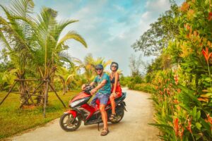 Two tourists on a rental scooter posing on a quiet tropical road in Phu Quoc, helmets on, surrounded by palm trees and colourful plants