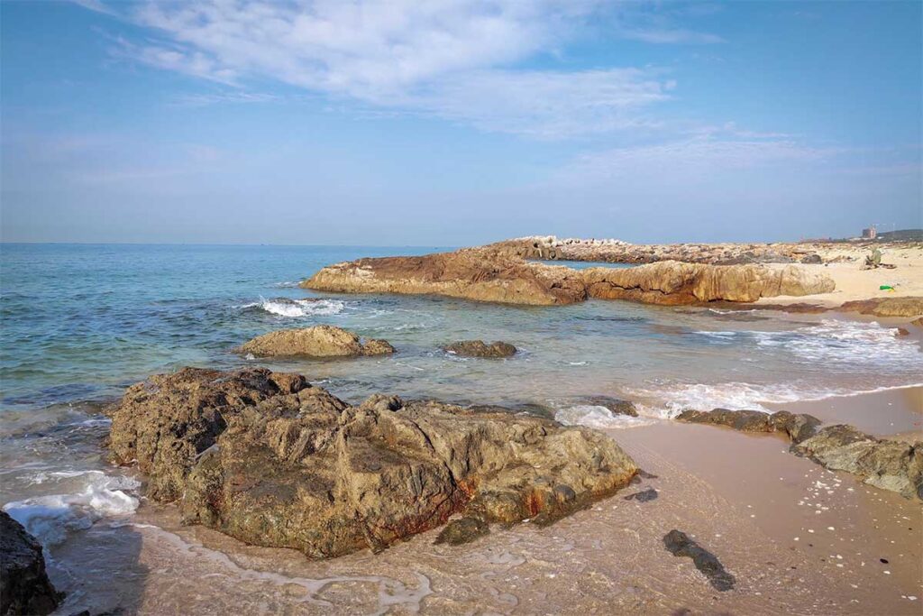 Rocky shoreline at Ong Dia Beach in Mui Ne, with large weathered rocks emerging from the sand and calm shallow water washing around them under a clear blue sky.