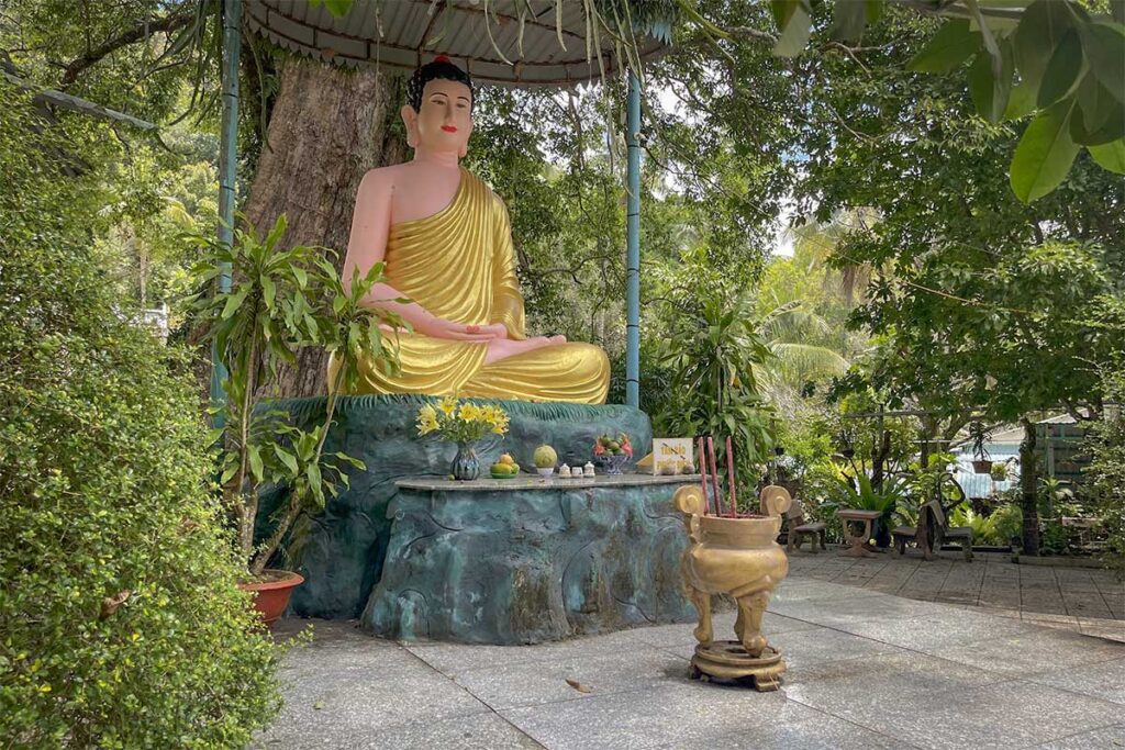 Seated Buddha statue under tree at Su Muon Pagoda Phu Quoc with incense burner and small offerings in peaceful garden area