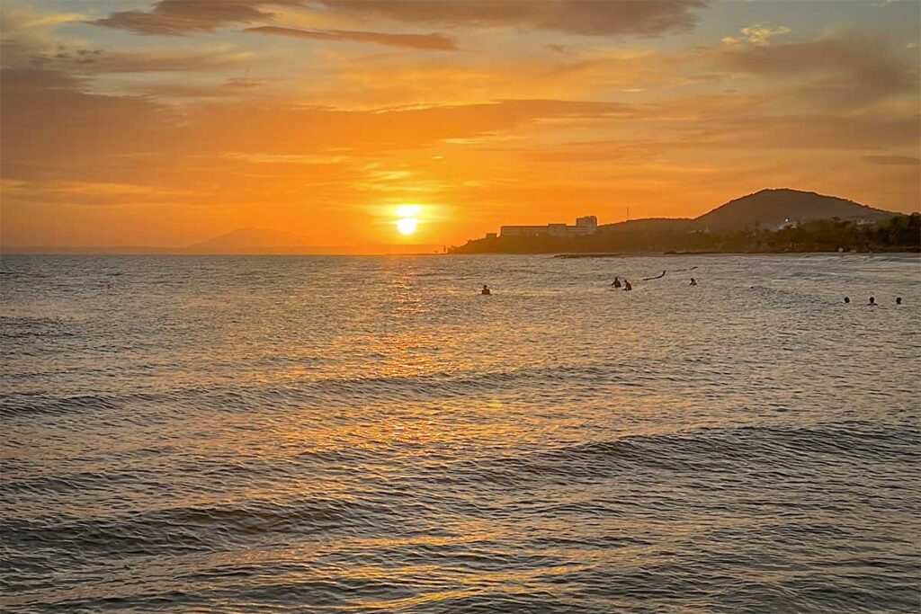 Golden sunrise over the sea at Ong Dia Beach in Mui Ne, with the sun setting low on the horizon and a group of surfers and swimmers silhouetted in the warm orange light on the water.