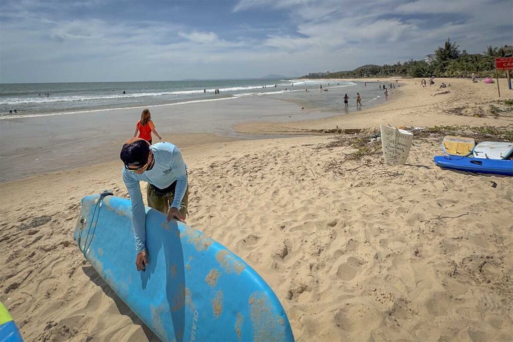 A surf instructor preparing a surfboard on the sandy beach at Ong Dia Beach in Mui Ne, with a handwritten surf school sign nearby and swimmers and surfers visible in the background.