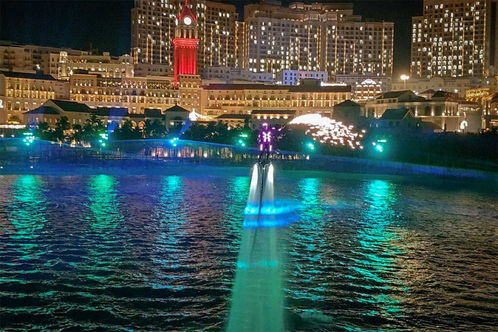 Flyboard performer glowing with blue lights rising above the sea during Symphony of the Sea Phu Quoc with bright waterfront buildings in the background