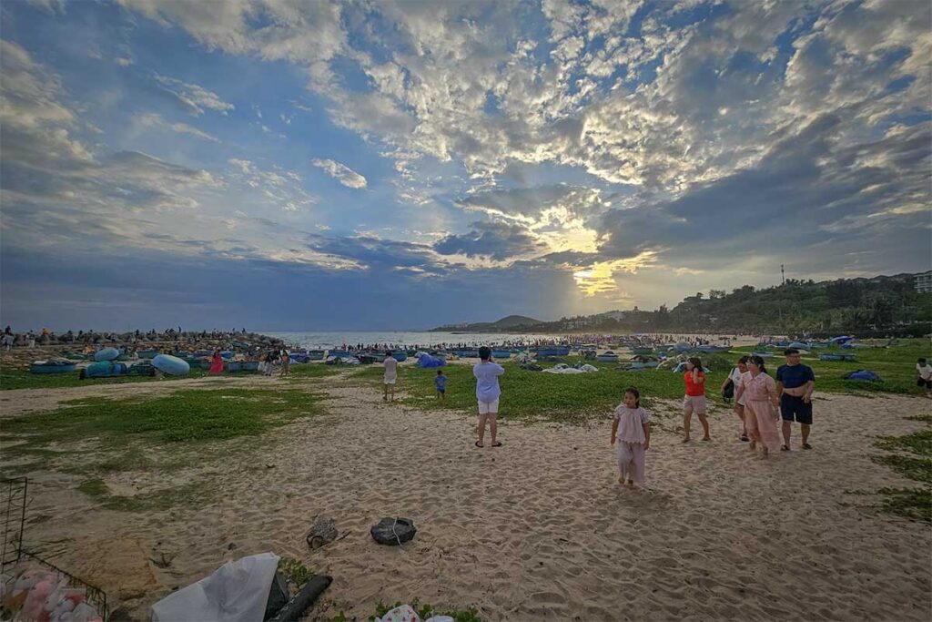 Locals and families gathering on Ong Dia Beach in Mui Ne during a busy late afternoon, with basket boats on the shore and a dramatic sunset sky overhead.