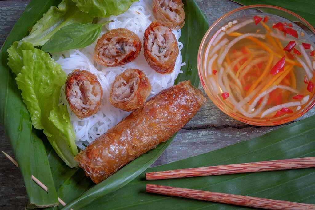 Vietnamese cha gio spring rolls served on a banana leaf with rice noodles, fresh lettuce, and a bowl of nuoc cham dipping sauce with chili and pickled vegetables.