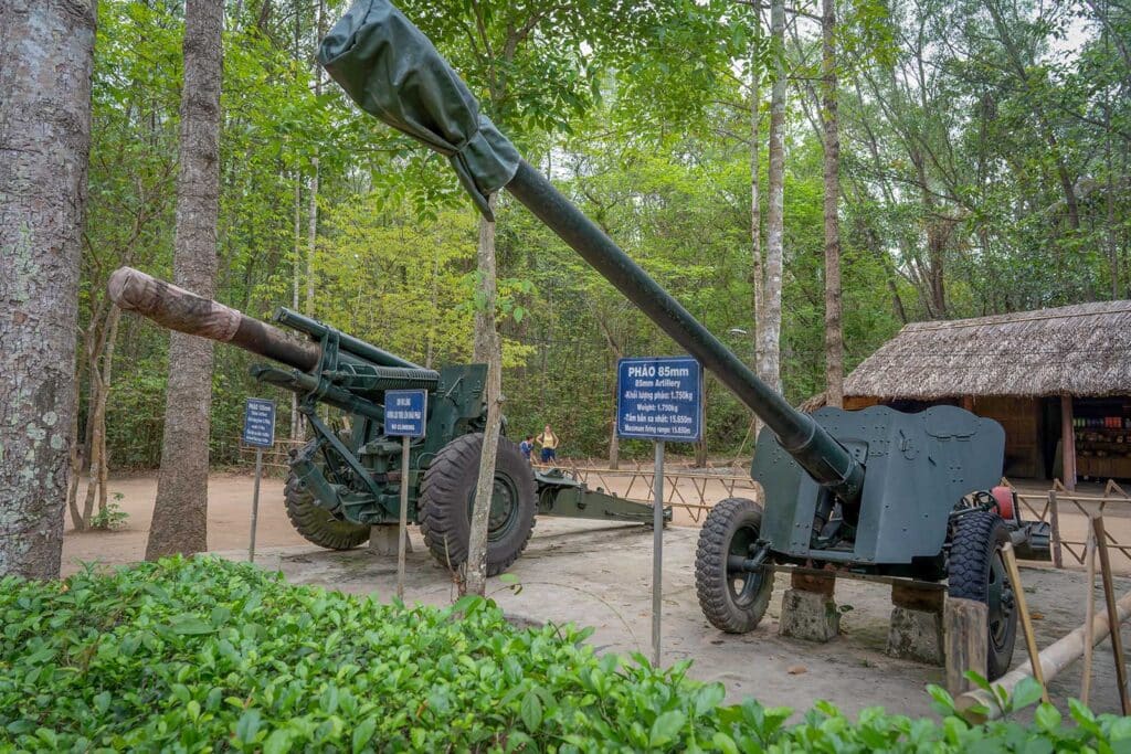 Two large artillery guns on display in the forest at the Cu Chi Tunnels near Ho Chi Minh City, with information signs and a thatched-roof shelter among the trees