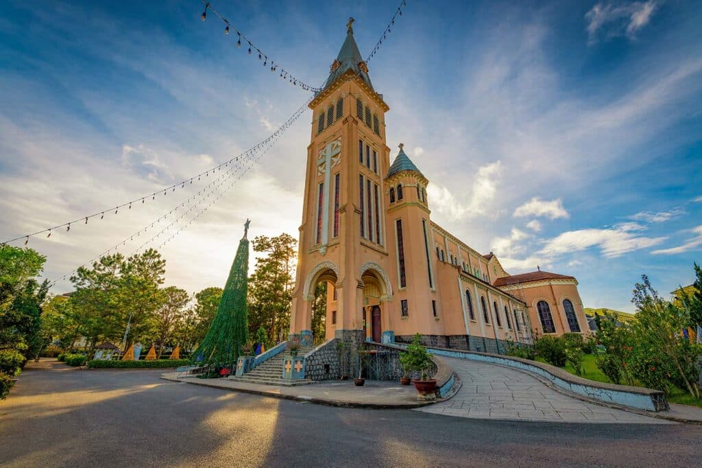 Dalat Cathedral, the pink church known as the chicken church, with its tall bell tower and spire under a blue sky, a string of lights and a cone-shaped topiary tree beside the entrance