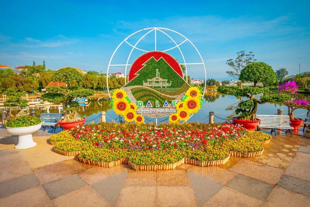 A large circular Dalat emblem made of flowers with sunflowers and a green globe frame, standing behind colorful flower beds and bonsai trees beside a calm lake at the Dalat flower garden