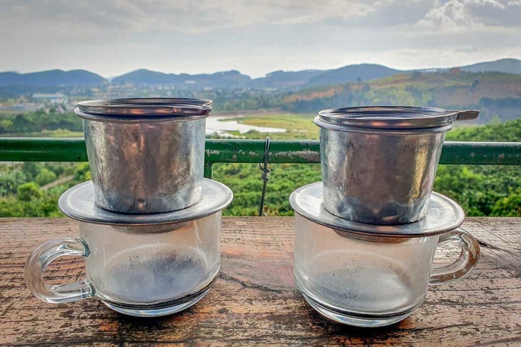 Two Vietnamese phin coffee filters brewing on a wooden railing at a hillside cafe in Dalat, with a view over green fields, a winding river, and misty hills in the distance