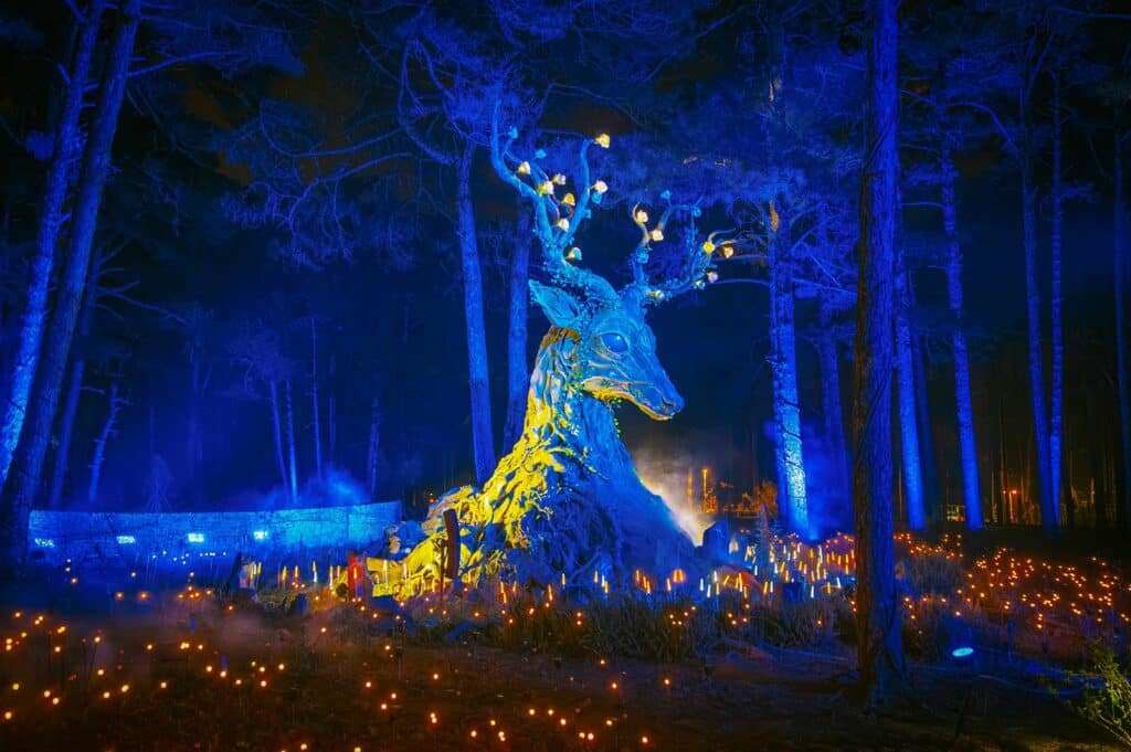 A giant illuminated deer sculpture glowing blue and yellow among tall pine trees at night, surrounded by a field of small warm lights, at a light park in Dalat