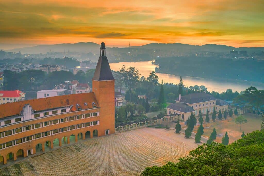 Elevated view of the Dalat Pedagogical College with its brick bell tower and orange-tiled roof at sunset, overlooking the town and a winding lake under a glowing orange sky