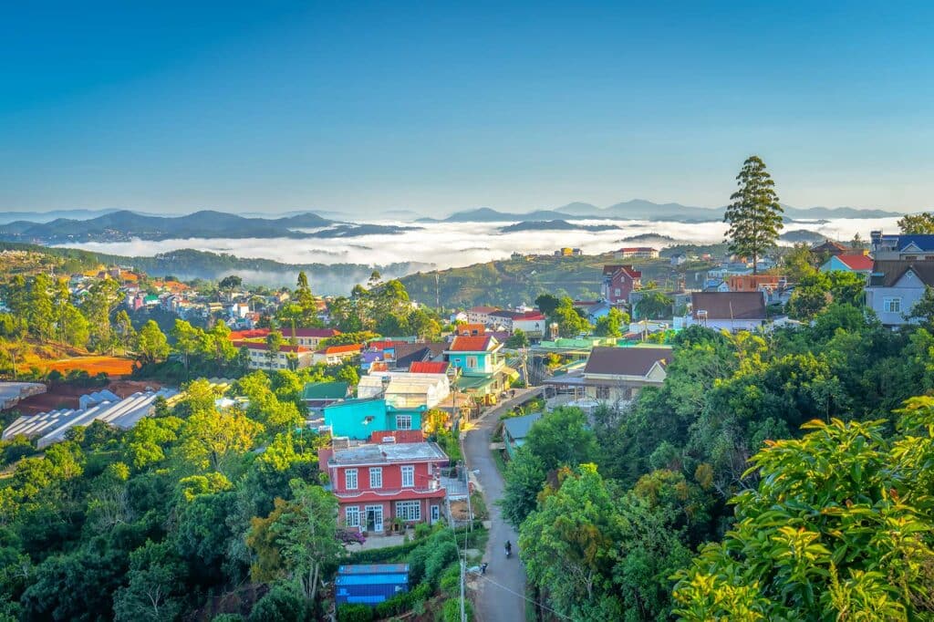Elevated view over Dalat town with colorful houses set among pine forest and greenhouses, morning mist rolling across the valley toward distant mountains